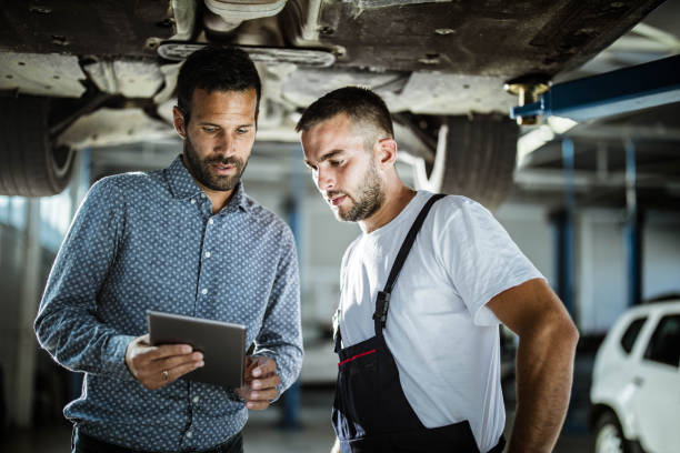 Manager and auto mechanic using touchpad in a workshop. Male customer and mechanic cooperating while using digital tablet in auto repair shop. auto repair shop mechanic digital tablet customer stock pictures, royalty-free photos & images