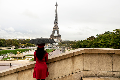 Young woman in a black dress is standing gracefully on old stairs near the Eiffel Tower in Paris and enjoying the atmosphere of the beautiful city. She seems greatly relaxed and elegant. Great composition with the Eiffel Tower in the background. Copy space available. Black and white image.