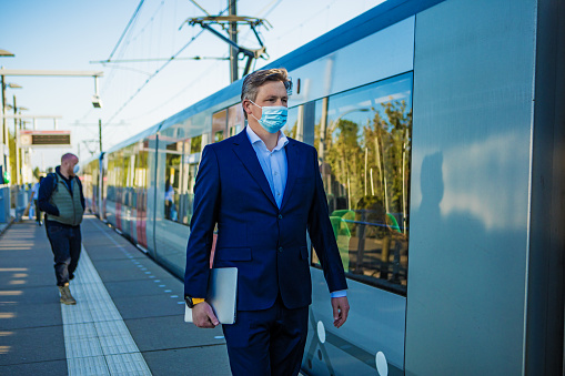 A handsome businessman wearing a suit waiting for public transport whilst wearing protective face masks at a train station during virus outbreak