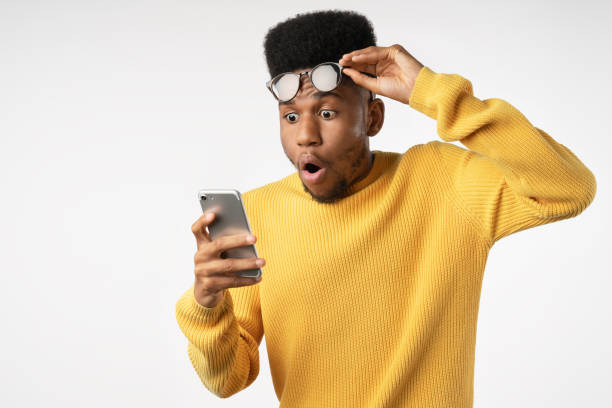 shocked african man while watching in smartphone on isolated white background - surprise imagens e fotografias de stock