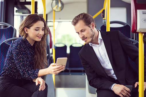 Young businesswoman using smart phone while traveling in public transport. Male professional is sitting by businesswoman. They watching mobile phone together.