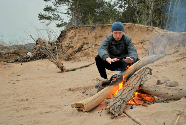 istock A man in a knitted hat, gray jacket and dark jeans sits on a spring evening by the fire on the sandy bank of the river and warms his hands. 1222977732