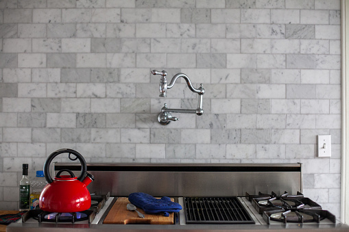 A pot filler faucet is a swing-out faucet on a long, jointed arm, mounted over the stove. Photograph features a pot filler faucet over a stove with a red tea kettle and a blue oven mitt and a gray brick wall.