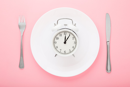 Reloj despertador en placa blanca con tenedor y cuchillo sobre fondo de mesa rosa claro. Color pastel. Closeup. Hora del almuerzo. Concepto de espera de comida. Vista de arriba hacia abajo. photo