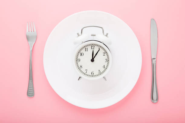 istock Alarm clock on white plate with fork and knife on light pink table background. Pastel color. Closeup. Lunch time. Meal waiting concept. Top down view. 1223000458