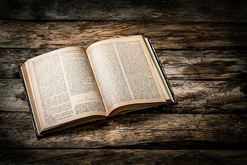 High angle view of an open old book with blurred text on pages shot on dark rustic wooden table. Useful copy space available for text and/or logo at the right of the book. Predominant color is brown. High resolution 42Mp studio digital capture taken with Sony A7rII and Sony FE 90mm f2.8 macro G OSS lens