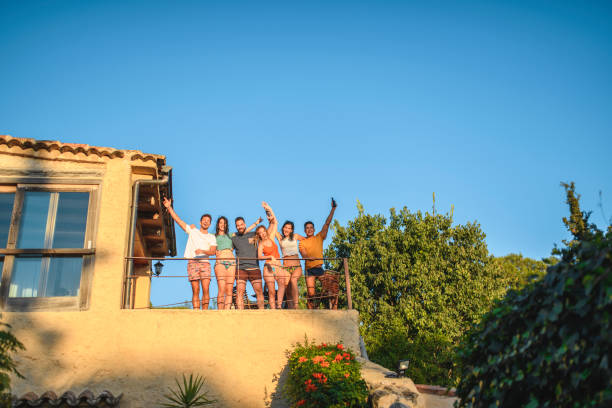 Male and Female Friends Waving from Farmhouse Terrace Low angle mid distance view of Spanish men and woman in 20s and 30s standing at farmhouse second floor terrace railing and waving at camera. male swimsuit standing arm around stock pictures, royalty-free photos & images