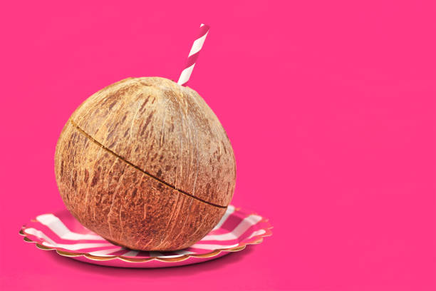 Whole Coconut fruit with drinking straw on  plate on left side of pink background with blank copy space on right side