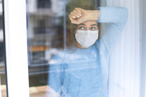 Mujer joven en cuarentena usando una máscara y mirando a través de la ventana photo