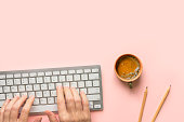 Hands of young Caucasian woman working typing on computer keyboard cup of hot coffee pencils on pink desktop.Home office freelance distant remote work