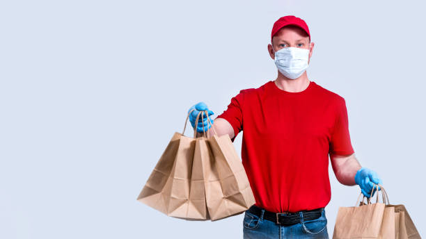 istock Banner. Safe delivery. A courier in a red uniform and protective mask and gloves holds a large order, many paper bags, contactless delivery food in quarantine. Donation of volunteers. Copy space. 1223023884