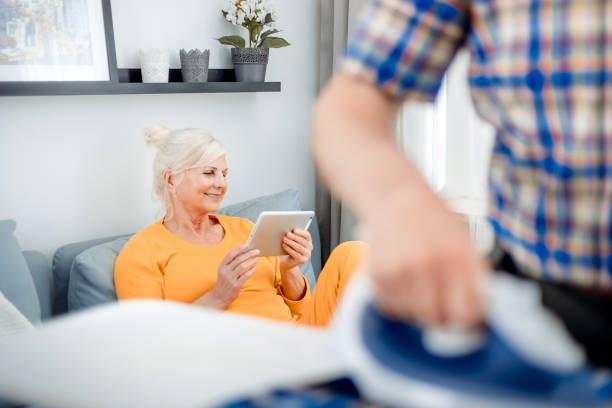 istock Senior wife using tablet while her husband doing housework with iron and clothes 1223026643