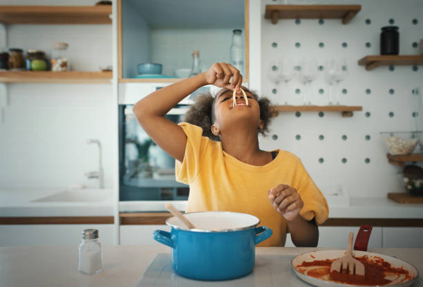 Little girl playing in the kitchen. Closeup side view of cute african american little girl playing in the kitchen and pretending to be a chef. She’s tasting pasta to see if it’s done. kids eating noodles stock pictures, royalty-free photos & images