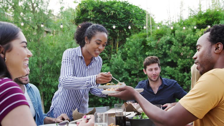 Woman Serving Group Of Multi-Cultural Friends At Home At Table Enjoying Food At Summer Garden Party