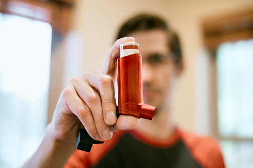 A young man at home uses his inhaler to help manage symptoms of asthma or cystic fibrosis.  Fitting imagery for National Asthma and Allergy Awareness Month.