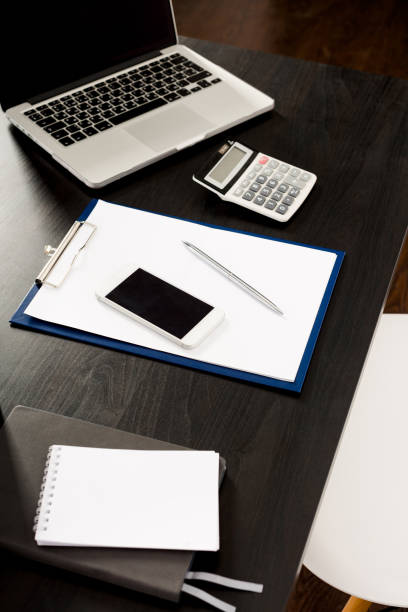 istock Laptop, smartphone, calculator, eyeglasses, pen, notebook, clipboard with blank white paper sheet and coffee on a black wooden desk, top view top view from the corner 1223036567