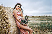 Young brunette in a pink satin dress, without linen standing near hay bales in windy weather. A woman holds a bouquet of wildflowers