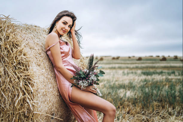 Young brunette in a pink satin dress, without linen standing near hay bales in windy weather. A woman holds a bouquet of wildflowers stock photo