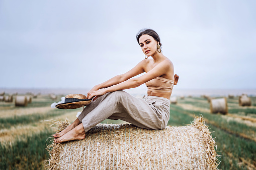 Barefoot brunette in linen pants and bare shoulders sitting on a hay bales in warm autumn day. Woman looking at camera. Behind her is a wheat field.