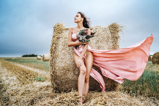 Young brunette in a pink satin dress, without linen standing near hay bales in windy weather. A woman holds a bouquet of wildflowers.