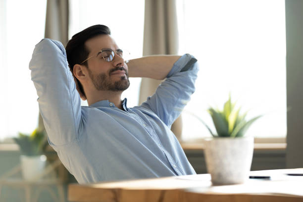 Peaceful young man daydreaming, leaning back, sitting at work desk Peaceful young man wearing glasses daydreaming with closed eyes, lazy sleepy businessman or student leaning back in comfortable chair, stretching hands, sitting at work desk, dreaming and visualizing man sleeping chair stock pictures, royalty-free photos & images