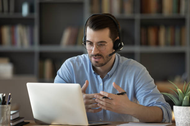 Confident man teacher wearing headset speaking, holding online lesson Confident man teacher coach wearing headset speaking, holding online lesson, focused student wearing glasses looking at laptop screen, studying, watching webinar training, listening to lecture phone speak stock pictures, royalty-free photos & images