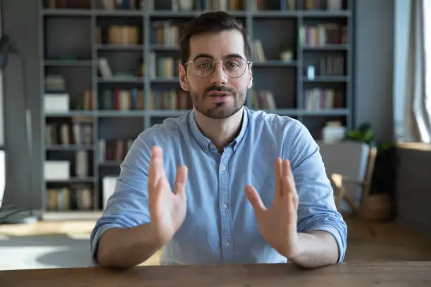 Head shot portrait confident businessman coach wearing glasses looking at camera and talking, mentor speaker holding online lesson, explaining, sitting at wooden work desk in modern cabinet