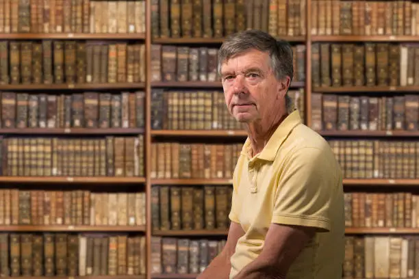 Photo of Senior caucasian man sitting in library full of old books on shelves