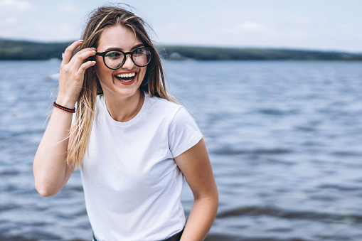 Portrait of a beautiful young woman in stylish glasses. Girl in white tshirt posing on the background of the lake landscape.