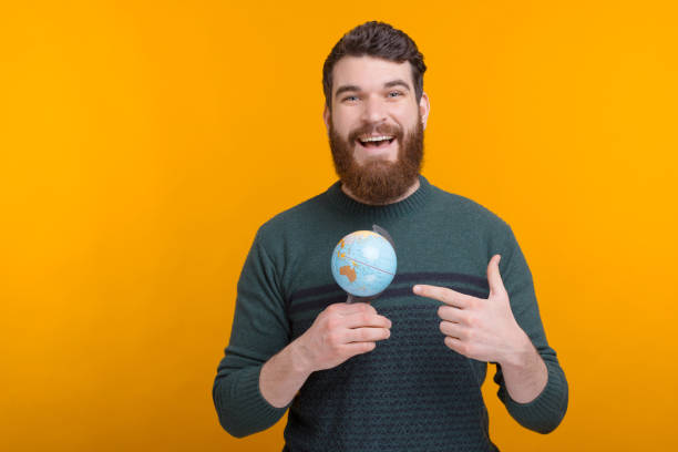 un joven estudiante masculino está apuntando a su pequeño globo sobre el fondo amarillo. - tourist map men holding fotografías e imágenes de stock
