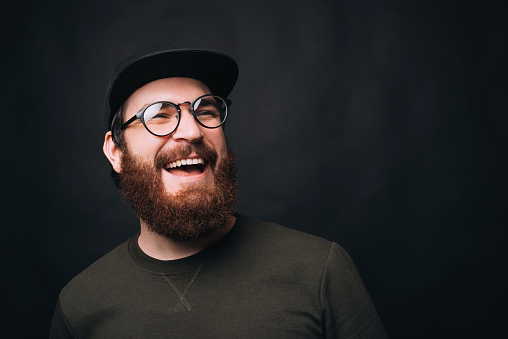 Close up photo of a bearded man wearing cap and glasses with a large smile on black background.