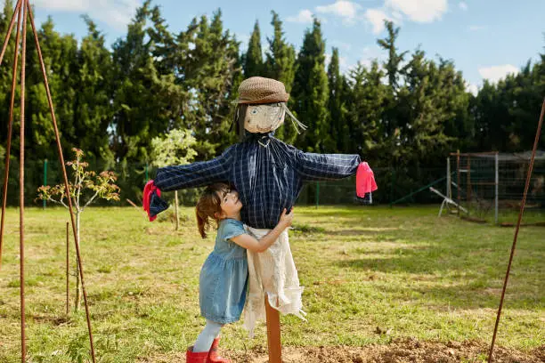 Enthusiastic 4 year old girl wearing denim dress and rubber boots smiling as she embraces scarecrow in community garden.