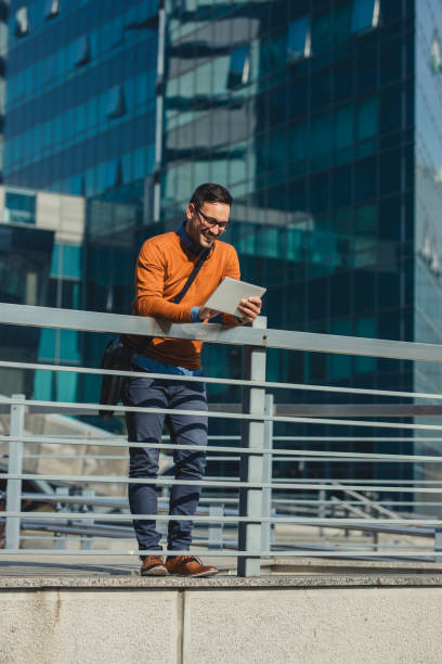 Successful Modern Young Businessman Using a Digital Tablet on the Street Handsome man standing in front of an office building and working remotely on his tablet. business person one man only blue standing stock pictures, royalty-free photos & images