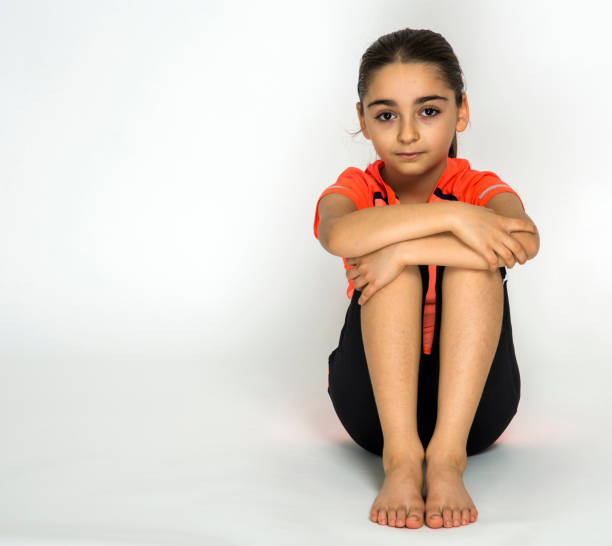 Studio photo of young girl seated on floor hugging knees on white background.