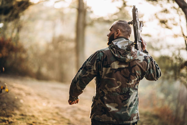 A camouflage soldier playing airsoft outdoors in the forest, sunset time, aiming at the rifle, back view stock photo