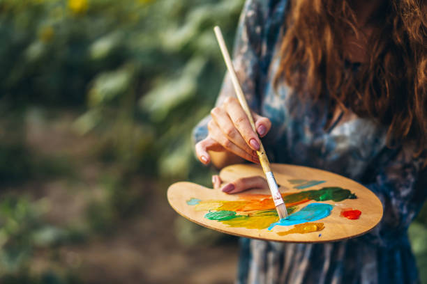 chiudi le mani dell'artista femminile tenendo pennello e tavolozza con vernici ad olio. sfondo sfocato con cavalletto in campo girasole - painting artist landscape painted image foto e immagini stock