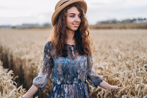 Closeup portrait of a beautiful young woman with curly hair. Woman in dress and hat standing in wheat field.