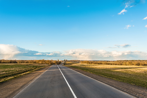 Picture of empty countryside road, horizontal image of a long straight empty highway flanked by green and yellow fields, blue sky with rare clouds in spring time