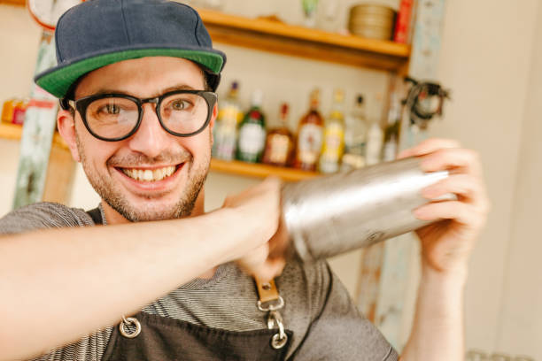 cameriere che indossa un grembiule, occhiali e un berretto sorridente mentre scuote un cocktail - shakerismo foto e immagini stock