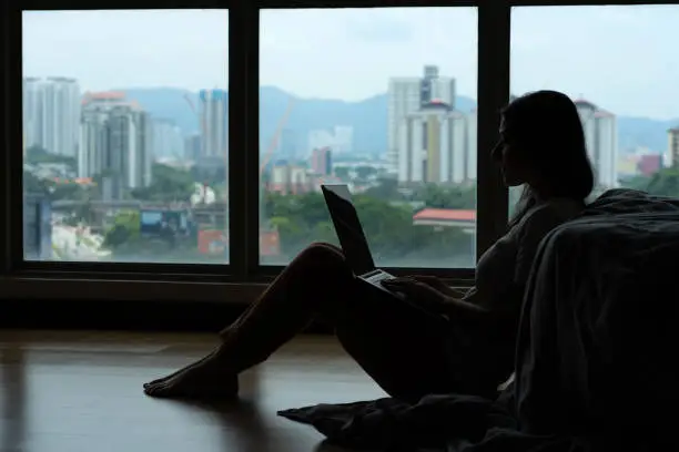 Siluet girl working on a laptop and drinking coffee, sitting on the floor near the bed by the panoramic window with a beautiful view from the high floor. Stylish modern interior. Shopping Internet