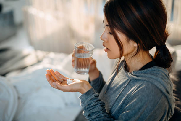 Young Asian woman sitting on bed and feeling sick, taking medicines in hand with a glass of water Young Asian woman sitting on bed and feeling sick, taking medicines in hand with a glass of water vitamin photos stock pictures, royalty-free photos & images