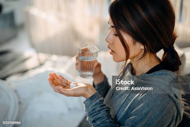 Young Asian Woman Sitting On Bed And Feeling Sick Taking Medicines In Hand With A Glass Of Water Stock Photo - Download Image Now