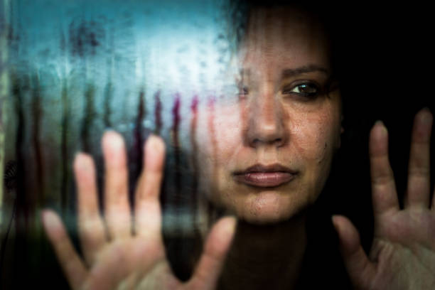 Depressed woman looking out of rainy window Close up desaturated color image depicting a sad, depressed-looking woman in her 30s, and of caucasian ethnicity, looking of a rain-soaked window with her hands pressed to the glass. The image was taken during the Covid-19 pandemic during lockdown, and the image illustrates a mental health concept, as well as a domestic abuse concept. Room for copy space. trap stock pictures, royalty-free photos & images