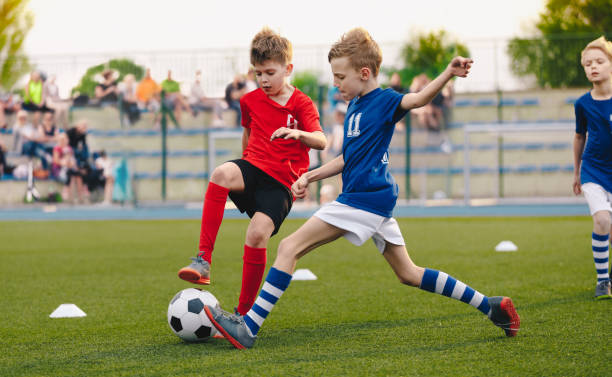 kids football players kicking ball on soccer field. sports soccer horizontal background. spectators on stadium in the background. youth junior athletes in red and blue soccer shirts. sports education - soccer ball team sport stadium soccer field imagens e fotografias de stock