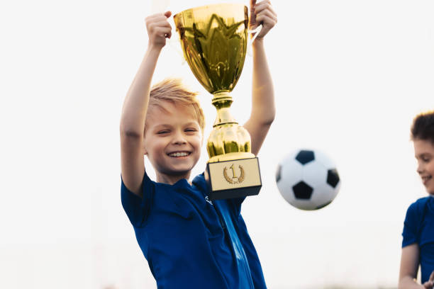 ragazzo felice che alza trofeo d'oro. competizione sportiva vincente per bambini. ragazzo felicissimo che detiene il premio d'oro. pallone da calcio sullo sfondo - medal soccer success winning foto e immagini stock