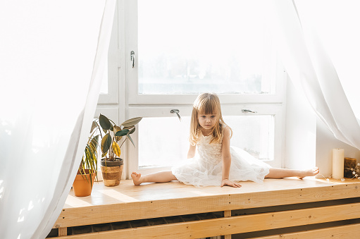 Cute little girl in beautiful dress is drop into the splits on a window sill at light sunny bedroom