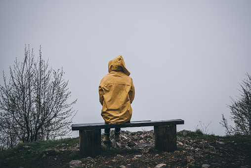 Rear view of depressed woman sitting alone on bench in yellow raincoat