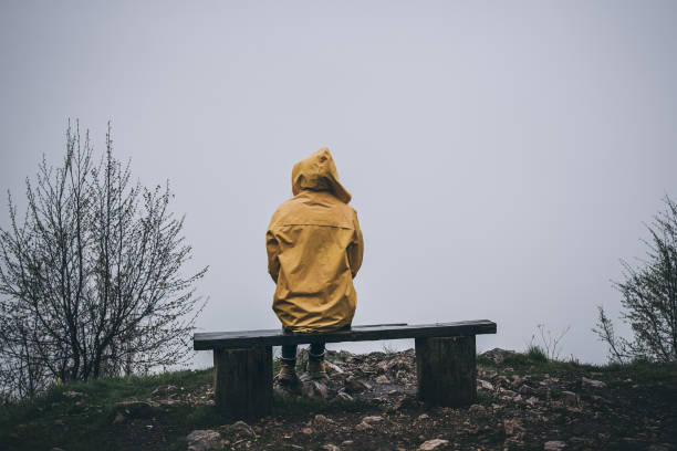Depression Rear view of depressed woman sitting alone on bench in yellow raincoat struggle stock pictures, royalty-free photos & images