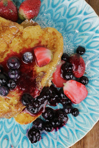 Stock photo of homemade eggy bread often called French toast on patterned, turquoise plate. This breakfast has been served with strawberries and blueberries and sprinkled with granulated sugar and fruit juice.