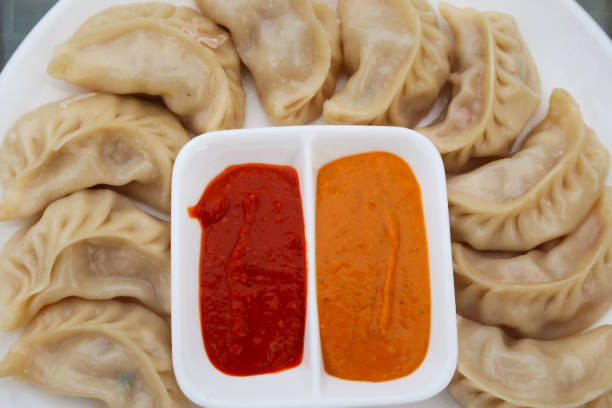 Image of steamed Momos (South Asian dumplings), white flour and water dough filled with chicken and mixed vegetables on plate with red and orange spicy dipping sauce, elevated view Stock photo showing a restaurant meal of steaming dumplings (Momos) filled with mixed vegetables and chicken on plate with red and orange spicy dipping sauce. chinese dumpling stock pictures, royalty-free photos & images
